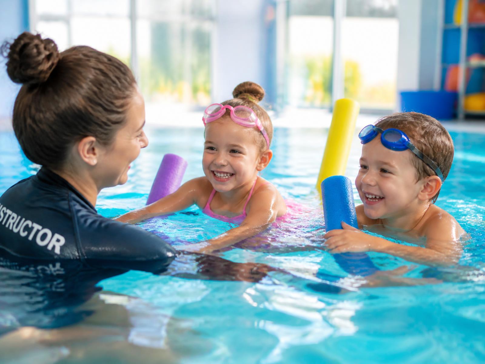 Swim instructor teaching two smiling young children in an indoor pool